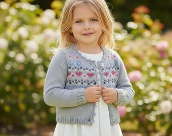 Puede incluir: Un cárdigan gris claro con un patrón de corazones y flores. El suéter de punto tiene cuello redondo y cierre de botones. El diseño incluye detalles en rosa, azul y blanco. La niña lleva un vestido blanco.