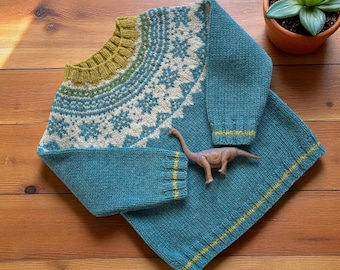 Puede incluir: Un suéter tejido y una manta a juego de color turquesa, con un patrón de copo de nieve en la pechera. El suéter tiene una pechera verde y crema y ribetes amarillos. Un juguete de dinosaurio descansa sobre la manta.
