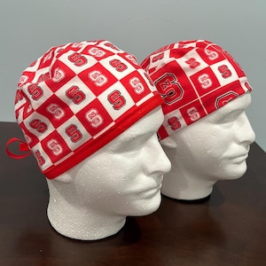 May include: Two red and white patterned scrub caps. The caps feature a repeating design of red squares with the letters "SC" and "OZ" in white. The caps are displayed on white mannequin heads.