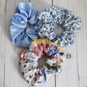 Puede incluir: Tres scrunchies de tela con patrones florales azules y blancos, uno con tela de sat&eacute;n azul claro s&oacute;lido.