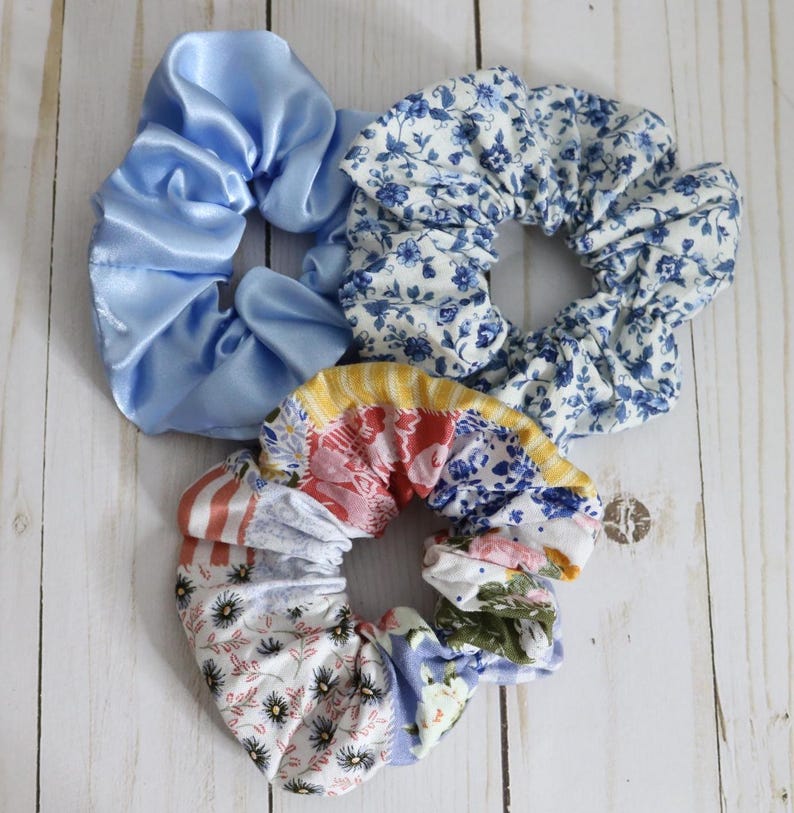 Puede incluir: Tres scrunchies de tela con patrones florales azules y blancos, uno con tela de sat&eacute;n azul claro s&oacute;lido.