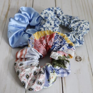 Puede incluir: Tres scrunchies de tela. Uno es de sat&eacute;n azul claro liso, otro es de estampado floral azul y blanco, y el tercero es un patchwork de varios estampados florales en azul, rosa, rojo y amarillo.