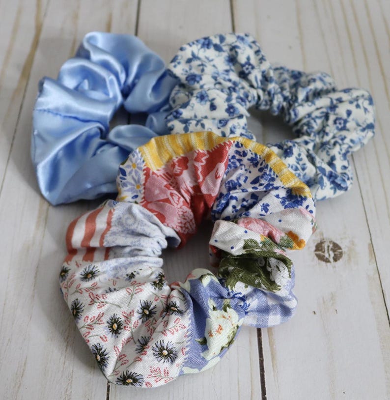 Puede incluir: Tres scrunchies de tela. Uno es de sat&eacute;n azul claro liso, otro es de estampado floral azul y blanco, y el tercero es un patchwork de varios estampados florales en azul, rosa, rojo y amarillo.