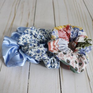 Puede incluir: Tres scrunchies de tela. Uno es de sat&eacute;n azul claro, otro es blanco con estampado floral azul y el &uacute;ltimo es un patchwork de varios estampados florales en rosa, azul y verde.