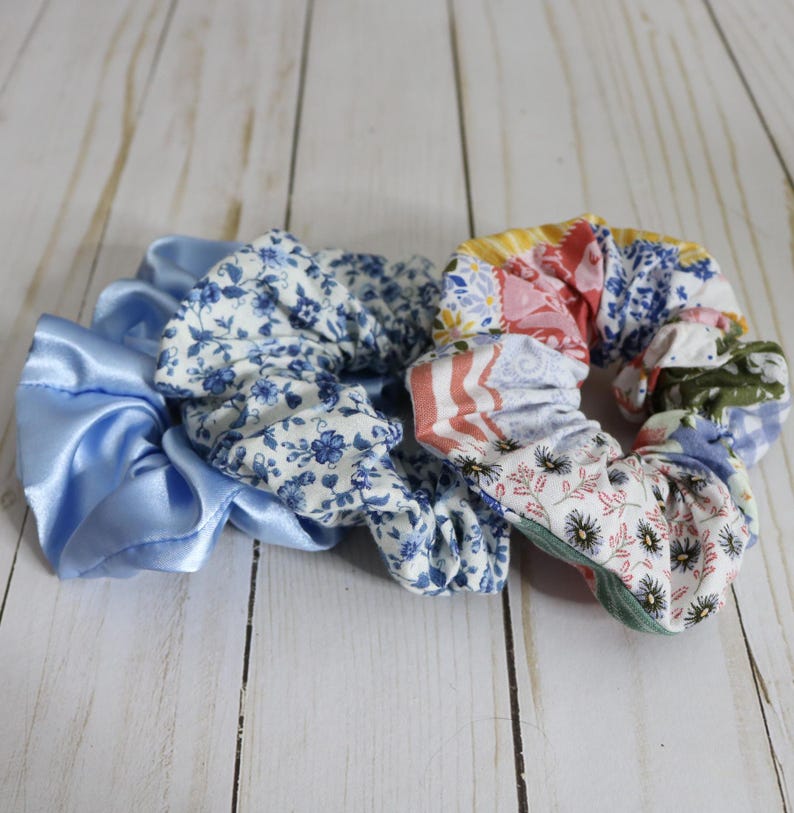 Puede incluir: Tres scrunchies de tela. Uno es de sat&eacute;n azul claro, otro es blanco con estampado floral azul y el &uacute;ltimo es un patchwork de varios estampados florales en rosa, azul y verde.