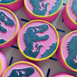 May include: Pink bath bombs with a blue dinosaur silhouette design. The bath bombs have a yellow rim.