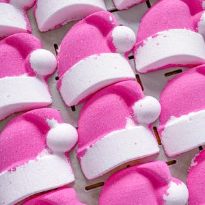 Peut inclure: Gros plan sur plusieurs bombes de bain roses et blanches en forme de bonnets de Père Noël. Les bombes de bain sont disposées en rangées sur une surface blanche.
