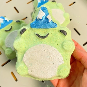 May include: Green bath bomb shaped like a frog wearing a blue and white hat. The frog has a white belly and black eyes.