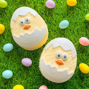 May include: Two yellow chick bath bombs with white cracked egg shells. The bath bombs are on a green grass surface with colourful Easter eggs scattered around.