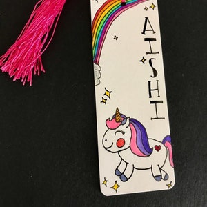 May include: A white bookmark with a rainbow, a unicorn, and the name "AISHI" written vertically. The bookmark has a pink tassel.