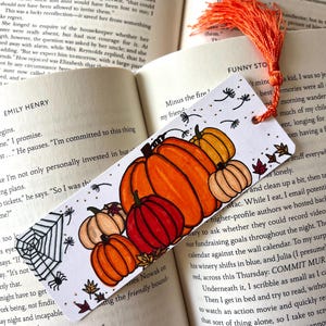 May include: A handmade bookmark featuring a colorful autumn pumpkin design. The bookmark has an orange tassel and is placed on an open book. The pumpkins are orange, red, and yellow with green leaves and a spider web.