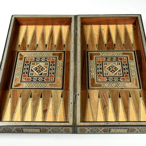 Peut inclure: Plateau de backgammon ouvert avec un design en bois incrusté complexe. Le plateau présente un cadre marron foncé, des triangles en bois clair et foncé et des incrustations carrées centrales décoratives avec des motifs géométriques. Le plateau est ouvert, révélant la surface de jeu.