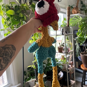May include: A handmade, plush chicken toy with a red head, white and black eyes, and a yellow beak. The body is teal and yellow, with long yellow legs. The toy is made of soft, textured yarn.