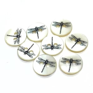 May include: Eight round, off-white buttons with a dragonfly design. Each button features a black dragonfly illustration. The buttons have two holes for sewing. They are arranged on a white surface.