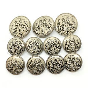 May include: A set of twelve silver-coloured metal buttons. Each button displays a detailed heraldic design with a castle, lions, and a shield. The buttons are round and have a slightly raised relief.