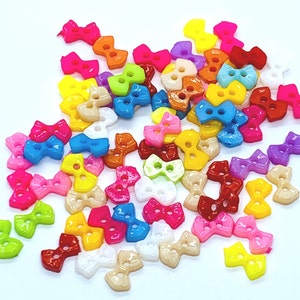 May include: Assortment of colourful plastic bow-shaped buttons. The buttons are in various colours, including pink, blue, yellow, green, red, and orange.
