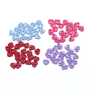 Peut inclure: Un assortiment de bloque-cordons en forme de cœur, en quatre couleurs : bleu clair, rose, rouge et lavande. Chaque bloque-cordon comporte deux trous pour l'insertion du cordon. Les bloque-cordons sont disposés en petits tas sur fond blanc.