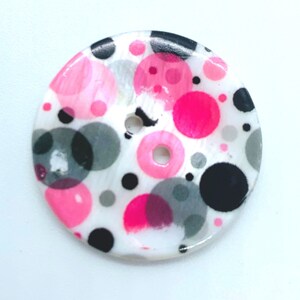 May include: A white button with a polka dot pattern in pink, black, and grey. The button has a two-hole design.
