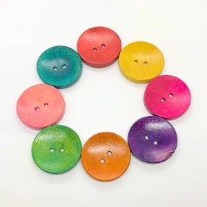 May include: A set of eight wooden buttons in various colours, including pink, orange, yellow, green, blue, and purple. Each button has two holes and a slightly rounded shape.