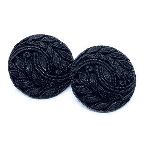 May include: A pair of round black buttons featuring a detailed embossed pattern of swirling leaves and vines. The surface of each button has a glossy finish with intricate textures that highlight the organic shapes of the foliage design.