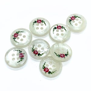 May include: Eight round, off-white buttons with a hand-painted design of a red rose and green leaves. Each button has four holes for sewing. The buttons are arranged on a white background, showcasing their floral design.