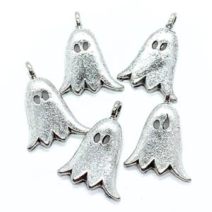 Op de afbeelding: Vijf zilverkleurige spookbedels met een getextureerd oppervlak. Elke bedel heeft twee ovale ooguitsparingen en een lus aan de bovenkant om aan sieraden of andere knutselwerken te bevestigen. De geesten hebben een golvende onderrand.