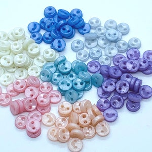 May include: Assortment of small, two-hole buttons in various pastel colours, including white, light blue, light purple, pink, and peach.