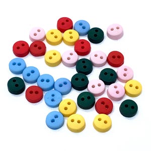 Peut inclure: Un assortiment de boutons ronds colorés, dans des tons de rouge, rose, jaune, bleu et vert. Chaque bouton comporte deux trous pour la couture. Les boutons sont dispersés sur une surface blanche.