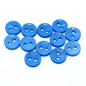 May include: A group of twelve light blue, round buttons, each with two holes. The buttons are arranged closely together and have a shiny, plastic appearance. The buttons are all the same shade of blue.