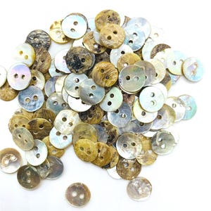 May include: A collection of round buttons with two holes, in shades of brown, tan, and iridescent white. The buttons have a natural, pearlescent sheen and are scattered against a white backdrop. Suitable for sewing and craft projects.
