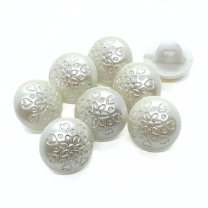 May include: A selection of off-white, round buttons with a pearl-like finish. Each button features a floral design with heart accents. One button is flipped over, revealing the back. These decorative buttons are suitable for various sewing and craft projects.