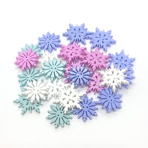May include: A collection of snowflake-shaped buttons in various pastel colours, including light blue, lavender, pink, and white. The buttons are made of plastic and have a smooth, glossy finish.