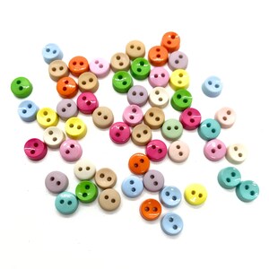 May include: A selection of small, round buttons in a variety of colours, such as pink, green, yellow, orange, and blue. Each button has two holes for sewing. The buttons are scattered on a white surface.