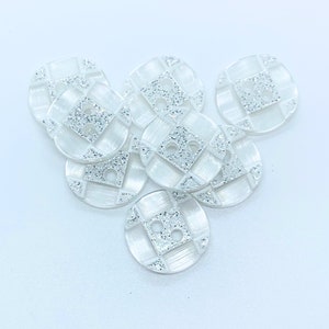 May include: A group of white round buttons with a silver glitter design and two holes. The buttons have a square pattern in the centre.