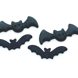 May include: Four black buttons shaped like bats with outstretched wings. Each bat button features two small holes in the centre for attachment. The buttons have a smooth, matte finish and are arranged in a scattered pattern on a plain background.
