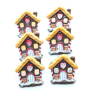May include: Six small gingerbread house buttons featuring brown walls, yellow windows, a light blue door, and a red and white candy cane trim. Each house has a textured yellow roof and a central red heart-shaped detail with a white cross pattern.