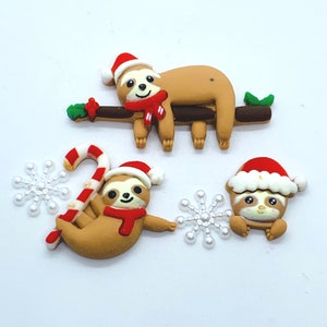 May include: Three Christmas-themed sloth ornaments, one on a branch, one holding a candy cane, and one sitting. Each sloth is wearing a Santa hat and a red scarf. The ornaments are surrounded by white snowflakes.