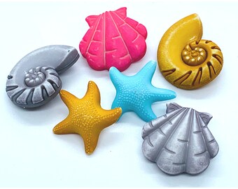 May include: Six colourful seashell-shaped buttons in various colours, including pink, gold, grey, blue, and yellow. The buttons are made of plastic and have a smooth, glossy finish.