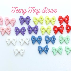 May include: A selection of miniature, brightly coloured bow-shaped decorations in shades of pink, purple, yellow, red, white, blue, and green. The text "Teeny Tiny Bows" is displayed in pink cursive font above the bows.