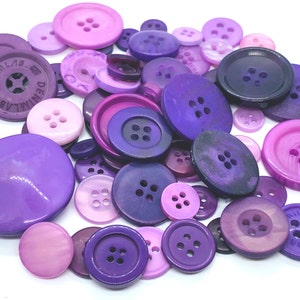 May include: A collection of purple and pink buttons in various sizes and styles. Some buttons have a shiny finish, while others have a matte finish. The buttons are arranged in a random pattern on a white background.