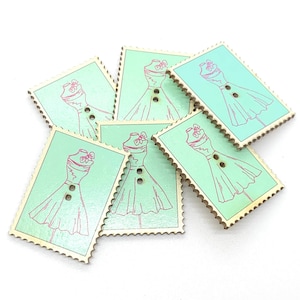 Op de afbeelding: Zes houten knopen in de vorm van postzegels met een groene achtergrond en een roze omtrek van een jurk met een strik op de halslijn.