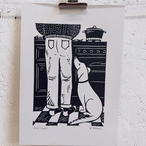 May include: Black and white linocut print showing a kitchen scene. A person in jeans and a sweater stands near an oven, with a dog looking up. The print includes the text "SOUS CHEF" and the artist's signature.