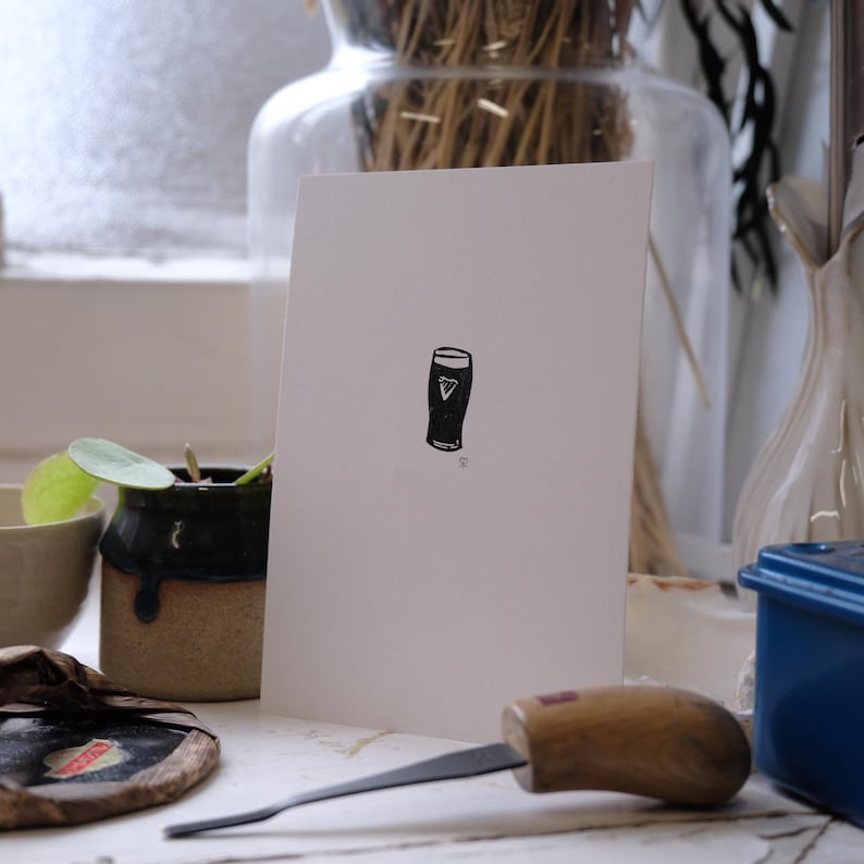 May include: A white greeting card features a black illustration of a pint of stout. The pint glass has a white triangle on the front. A small plant in a ceramic pot and a carving tool are in the foreground.