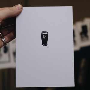 May include: A black and white print of a pint glass with a white background. The pint glass has a dark body and a white head, with a white triangle in the center. The print is held by a hand.
