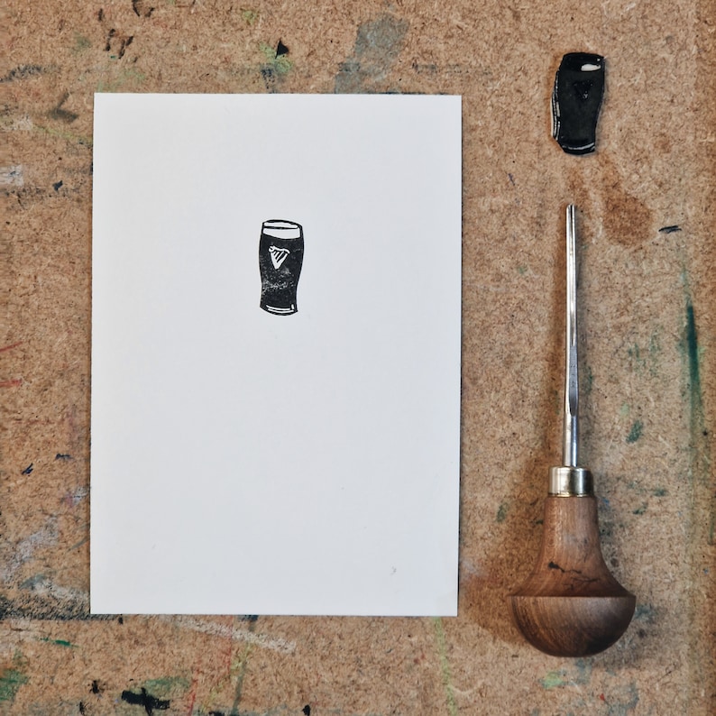 May include: A black and white print of a pint glass with the Guinness logo on a white paper. A black stamp of the pint glass and a carving tool are next to the print. The background is a wooden surface.