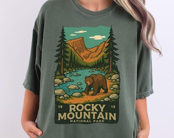 May include: Green t-shirt featuring a graphic of a bear walking near a river in a mountain landscape. The design includes trees, mountains, and the text "Rocky Mountain National Park" with the years "19" and "15".