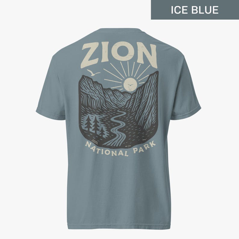 May include: A light blue t-shirt with a graphic print of a mountain landscape with a river, trees, and a sun. The text "Zion National Park" is printed below the graphic. The text "Zion" is printed above the graphic.