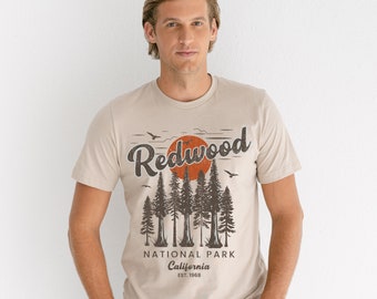 May include: A beige t-shirt with a vintage-style graphic of Redwood National Park, California. The graphic features a red sun setting behind a row of tall trees with the text "Redwood National Park California Est. 1968".