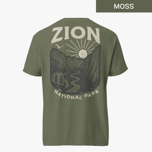 May include: A moss green t-shirt with a vintage-style graphic of Zion National Park. The graphic features a mountain range, a winding river, a sun, and a bird in flight. The text "Zion National Park" is printed below the graphic.