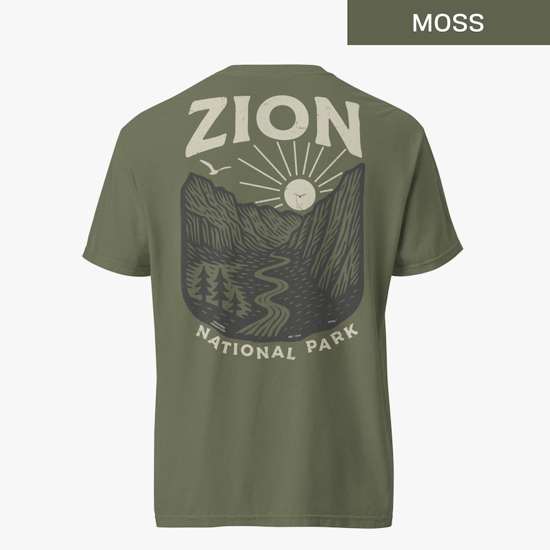 May include: A moss green t-shirt with a vintage-style graphic of Zion National Park. The graphic features a mountain range, a winding river, a sun, and a bird in flight. The text "Zion National Park" is printed below the graphic.